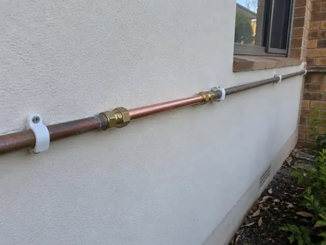 Repaired copper water pipe along an exterior rendered wall beside a brick window in Bennetts Green
